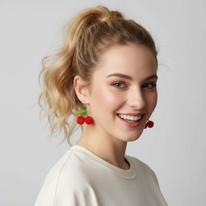 May include: A woman wearing red cherry earrings with green leaves. The earrings are a fun, playful accessory, perfect for adding a pop of color to any outfit. The earrings are a unique and eye-catching piece of jewelry.