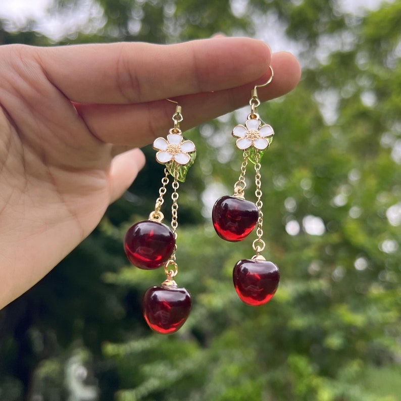 May include: Gold dangle earrings with a white flower charm and three red cherry charms.