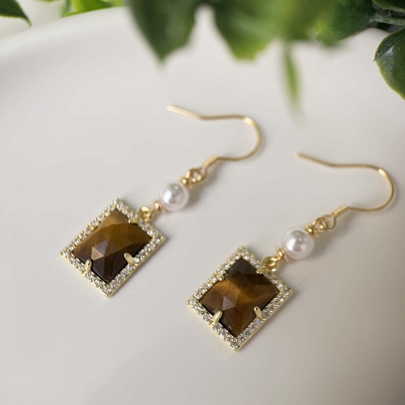 May include: Gold earrings with a square tiger's eye stone and a pearl accent.