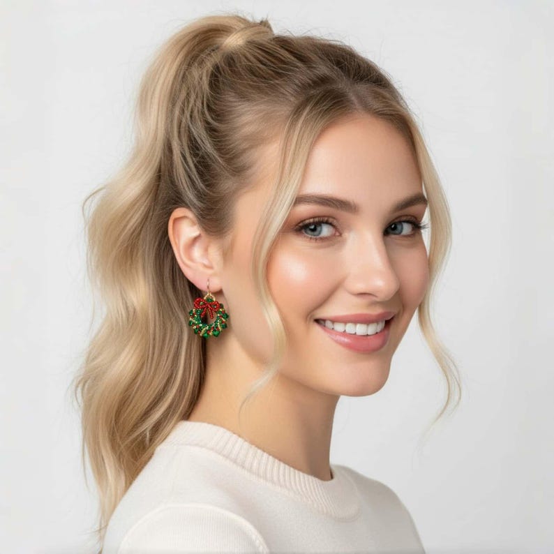 May include: Gold-colored Christmas wreath earrings with red bow accents. The earrings are green and red, with a festive design. The earrings are small and dangle from the ear.