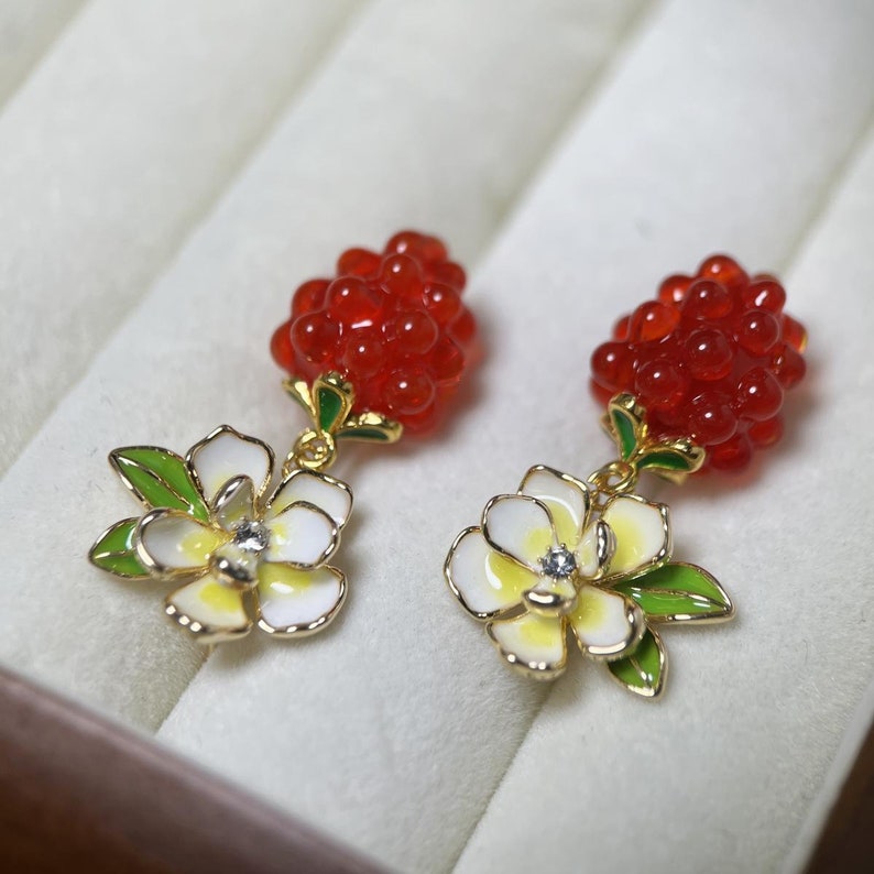 May include: A pair of dangle earrings with red berry clusters and white flower charms. The flowers have yellow centers and green leaves.