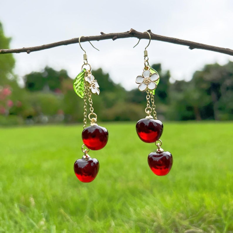 May include: Gold dangle earrings with a red cherry charm, a white flower charm, and a green leaf charm.