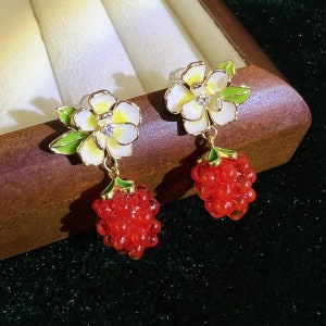 May include: A pair of gold earrings with white flower charms and red berry charms. The flowers have green leaves and a small rhinestone in the center.