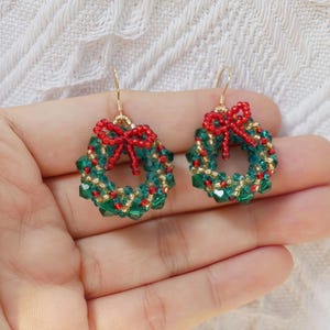 May include: A pair of Christmas wreath earrings. Each earring features a circular design made of green and gold beads, with a small red bow at the top. The earrings have gold-colored hooks.