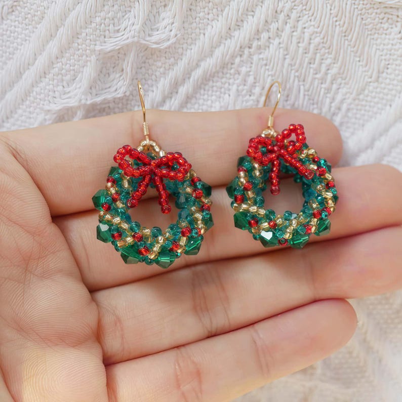 May include: A pair of Christmas wreath earrings. Each earring features a circular design made of green and gold beads, with a small red bow at the top. The earrings have gold-colored hooks.