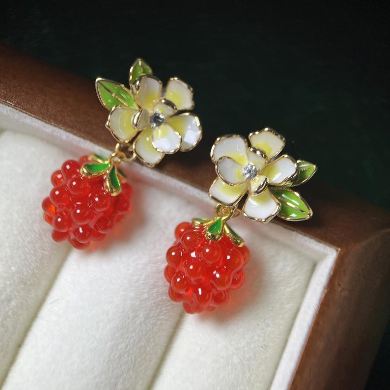 May include: A pair of dangle earrings with a white flower and red berry charm. The flower has a yellow center and green leaves. The berry is a cluster of small red beads.