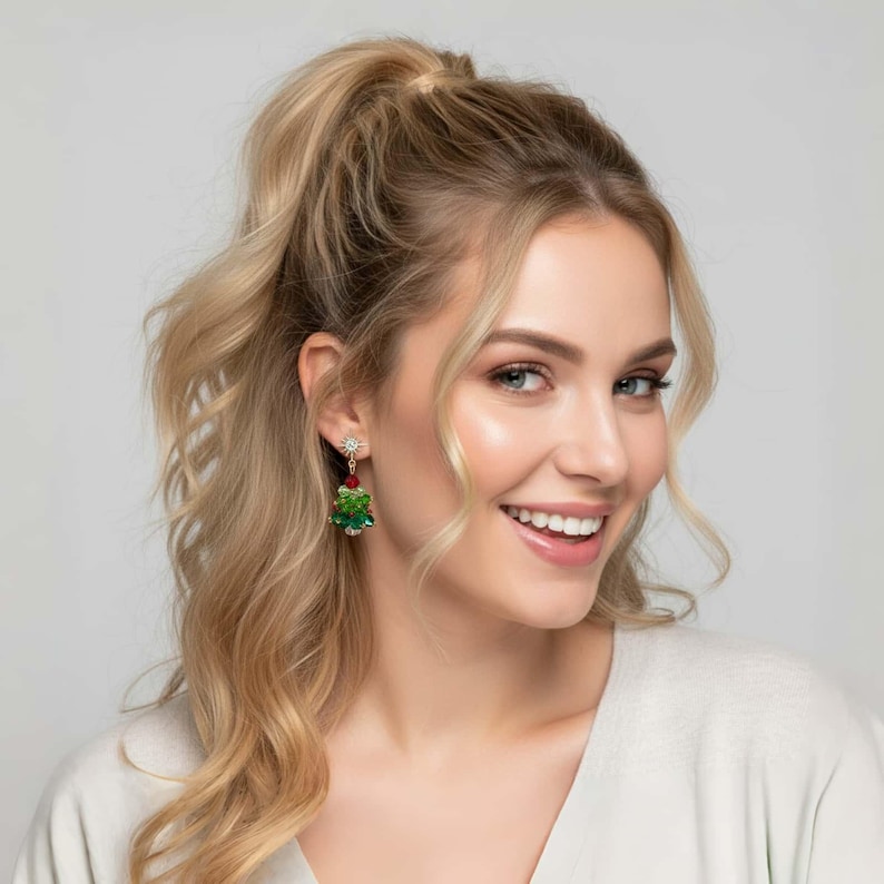 May include: Dangling Christmas tree earrings with red and green accents. The earrings feature a star-shaped stud with a red bead and a green, gem-studded tree design. The earrings are worn by a person with blonde hair.