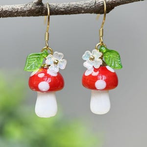 May include: A pair of gold dangle earrings featuring red and white mushroom charms with green leaves and white flowers.