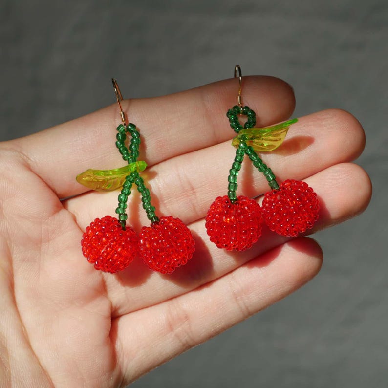 May include: A pair of beaded cherry earrings. Each earring features two red beaded cherries, green beaded stems, and a yellow leaf. The earrings have gold-colored ear wires.