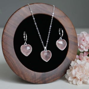 May include: A jewelry set consisting of a silver chain necklace and two matching hoop earrings, each featuring a faceted, heart-shaped light pink stone pendant. The pieces are displayed on a circular dark wood stand with a black velvet interior.