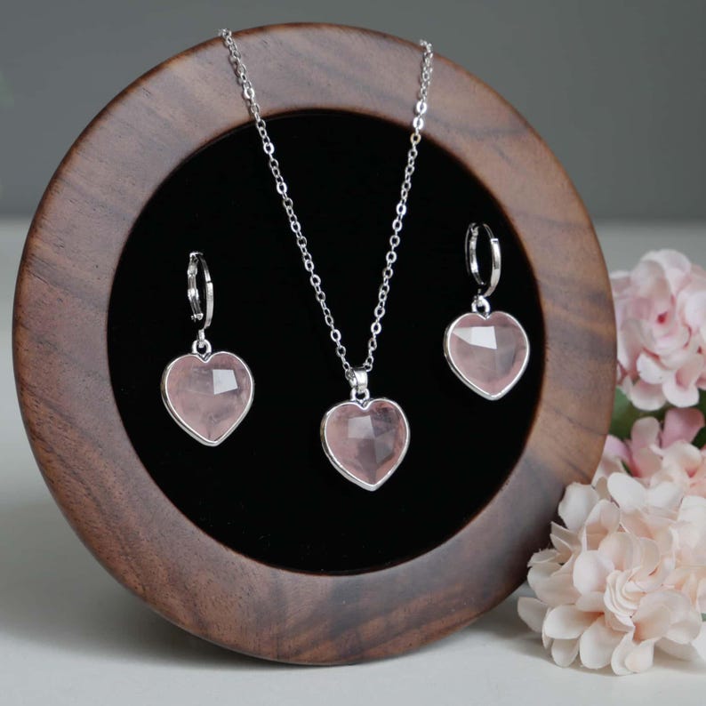 May include: A jewelry set consisting of a silver chain necklace and two matching hoop earrings, each featuring a faceted, heart-shaped light pink stone pendant. The pieces are displayed on a circular dark wood stand with a black velvet interior.
