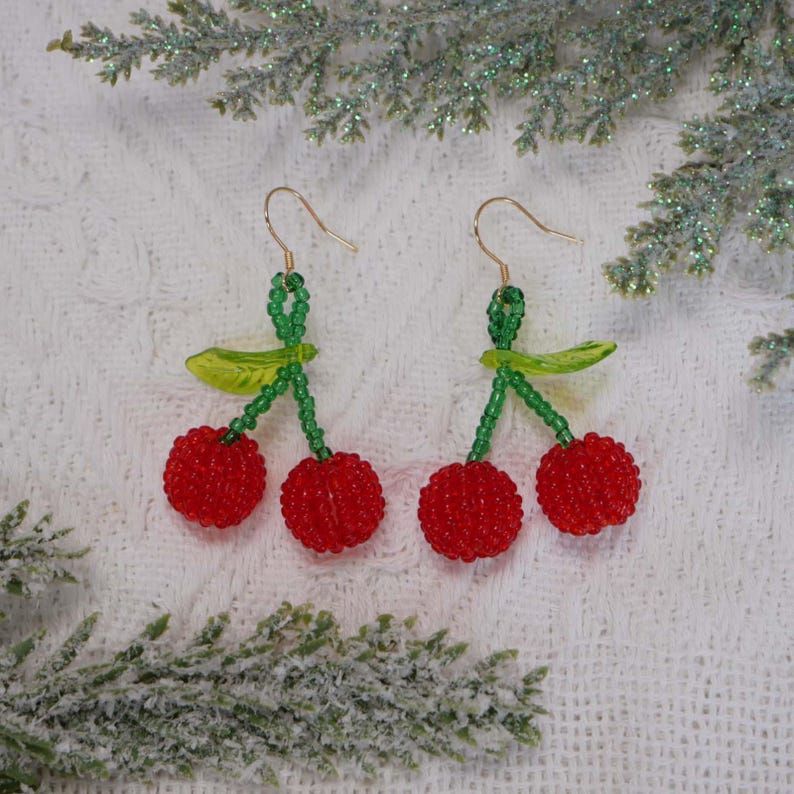 May include: A pair of cherry earrings with red beaded cherries, green beaded stems, and gold-colored ear wires. The earrings have a leaf detail and are set against a white background.