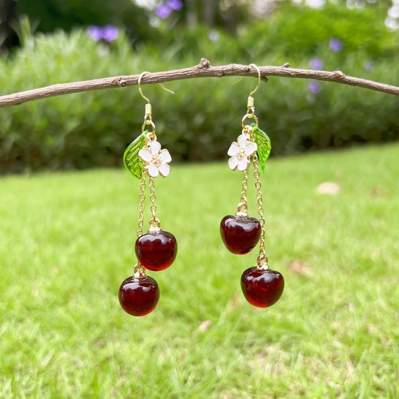 May include: Gold dangle earrings with a cherry and flower charm. The earrings feature three red cherries and a white flower with green leaves.
