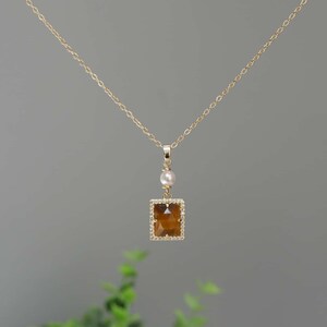 May include: A gold chain necklace with a square pendant featuring a brown tiger's eye stone surrounded by clear rhinestones. A single white pearl hangs above the stone.