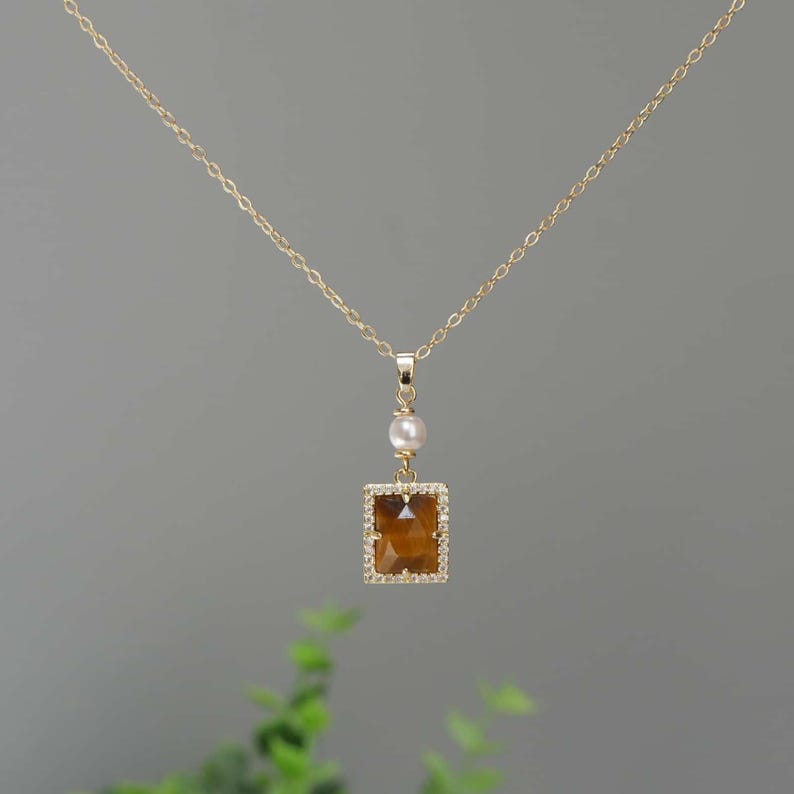 May include: A gold chain necklace with a square pendant featuring a brown tiger's eye stone surrounded by clear rhinestones. A single white pearl hangs above the stone.
