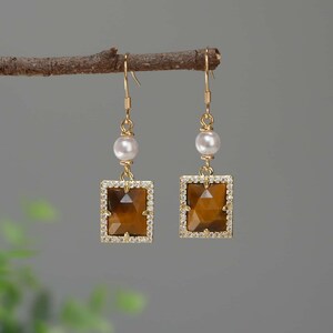 May include: Gold earrings with a brown tiger's eye stone set in a square frame with clear rhinestones. Each earring has a white pearl bead above the stone.