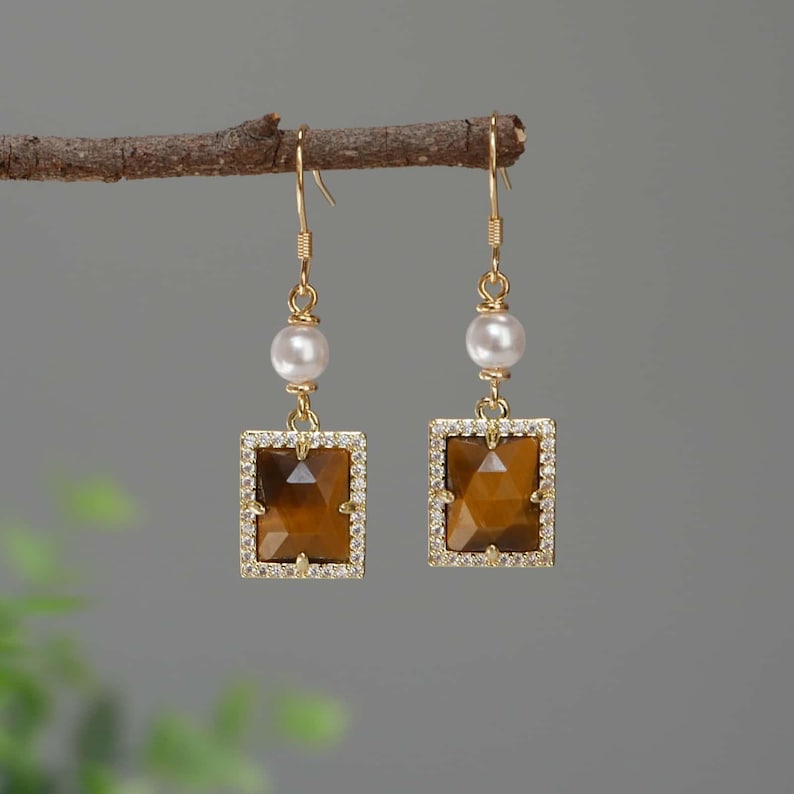 May include: Gold earrings with a brown tiger's eye stone set in a square frame with clear rhinestones. Each earring has a white pearl bead above the stone.