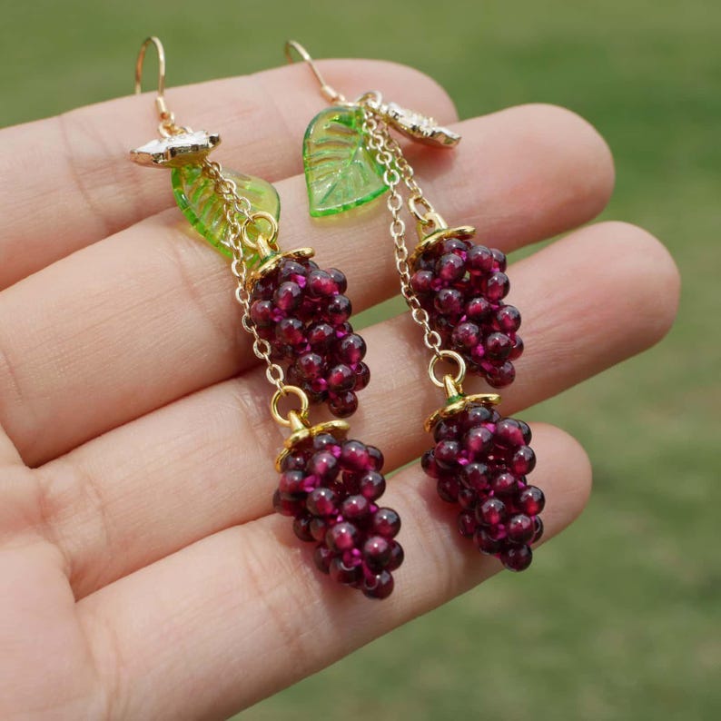 Op de afbeelding: Een paar oorhangers met twee trossen donkerrode granaatstenen, elke tros hangt aan een gouden ketting met een groene glazen bladeren charme.