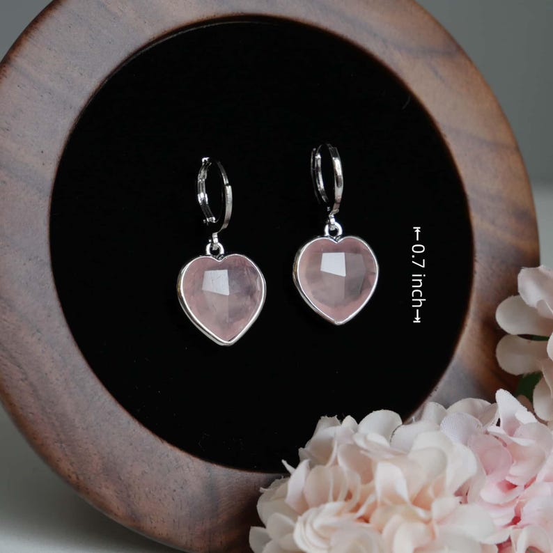 May include: A pair of silver-toned hoop earrings featuring faceted, heart-shaped light pink stone pendants. The earrings measure 0.7 inches in height as indicated by the text in the image. The design includes a secure hinged closure mechanism.