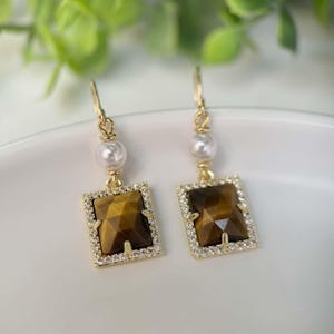 May include: Gold dangle earrings with a square tiger's eye gemstone surrounded by clear cubic zirconia stones and a white pearl accent.