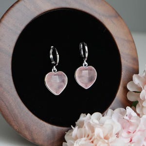May include: A pair of silver hoop earrings featuring faceted, heart-shaped light pink rose quartz gemstone drops. The earrings are displayed against a dark background within a circular wooden frame, highlighting the clear, polished stone texture.