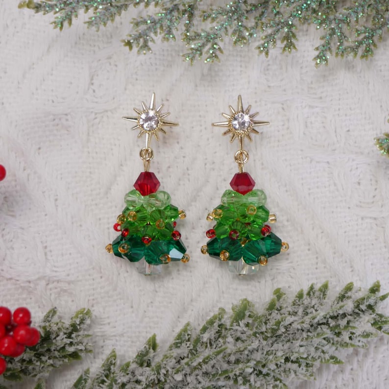 May include: A pair of Christmas tree earrings. Each earring features a gold starburst top, a red bead, and a green beaded tree with red accents. The earrings are set against a white background with decorative greenery.
