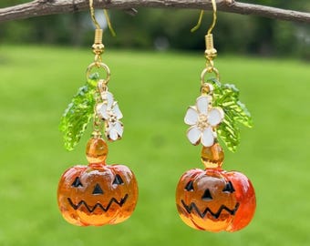 May include: A pair of orange pumpkin dangle earrings with white flowers and green leaves. The pumpkins have black jack-o-lantern faces.