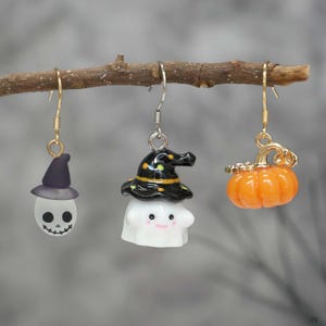 May include: Three Halloween-themed earrings suspended from a twig. The earrings feature a ghost with a witch's hat, a ghost with a skull face, and an orange pumpkin. The earrings have gold and silver hooks.