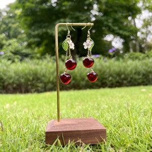 May include: Gold dangle earrings with a cherry design. Each earring features two red glass cherries, a green leaf, and a white flower.