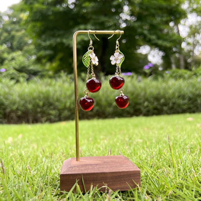 May include: Gold dangle earrings with a cherry design. Each earring features two red glass cherries, a green leaf, and a white flower.