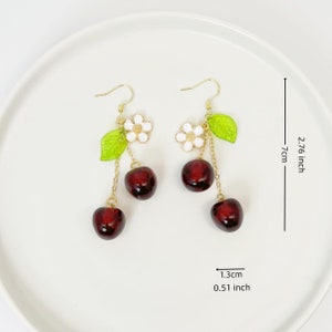 May include: Gold dangle earrings with three red cherry charms and a white flower charm with green leaves. The earrings are 2.76 inches long and the cherry charms are 0.51 inches wide.