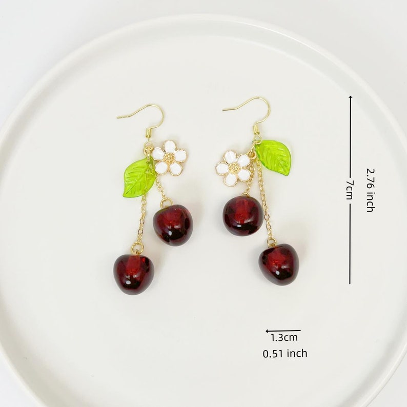 May include: Gold dangle earrings with three red cherry charms and a white flower charm with green leaves. The earrings are 2.76 inches long and the cherry charms are 0.51 inches wide.