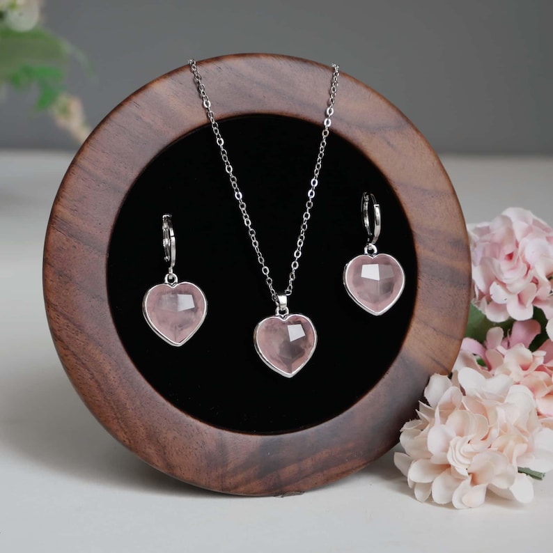 May include: A jewelry set featuring a silver-toned chain necklace and two matching hoop earrings, each adorned with a faceted, heart-shaped light pink stone pendant. The pieces are displayed on a circular dark wood stand with a black velvet center.