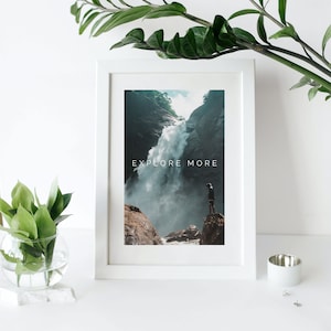 Peut inclure: Une impression encadr&eacute;e blanche avec une photographie d'une cascade dans une for&ecirc;t luxuriante et verte. Le texte "EXPLORE MORE" est imprim&eacute; en blanc sur l'image.