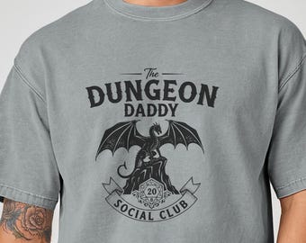 以下が含まれることがあります： 「The Dungeon Daddy Social Club」の文字とドラゴンのグラフィックがプリントされた、グレーの半袖Tシャツ。柔らかい素材でできており、ゆったりとしたフィット感です。「Granite」の文字が下部にプリントされています。