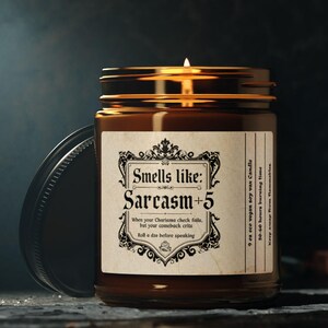 May include: A lit, brown glass jar candle with a black lid. The label reads "Smells like: Sarcasm + 5". The candle is on a dark surface. The candle's text also reads "When your Chartoons check fails, but your comeback crits, roll a die before speaking."