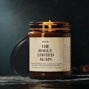 May include: A lit soy wax candle in a brown glass jar with a black lid. The label reads "Smells like THE ROGUE LOOTED AGAIN." The candle is on a dark surface with a blurred background.