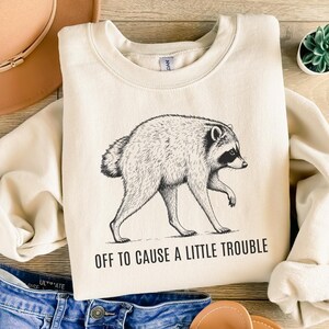 Puede incluir: Una sudadera de cuello redondo color crema con una ilustración de línea negra de un mapache caminando. Debajo del mapache, el texto dice "OFF TO CAUSE A LITTLE TROUBLE" en una fuente sans-serif simple y negrita. La sudadera tiene mangas largas.