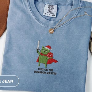 Könnte beinhalten: Hellblaues Comfort Colors T-Shirt mit einem gestickten grünen Kater mit Weihnachtsmütze und Umhang, der ein Schwert und einen Würfel hält. Der Text "Dibs on the Dungeon Master" steht darunter. Eine goldene Halskette liegt auf dem Shirt.