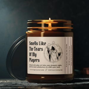 May include: A lit soy wax candle in a brown glass jar with a black lid. The label reads "Smells Like The Tears Of My Players" with a graphic of a cloaked figure. The candle is designed for 50-60 hours of burn time.