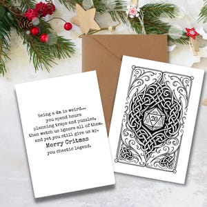 May include: Two white greeting cards with black text and illustrations, one with a dice design and the other with text that reads "Merry Critmas". A brown envelope is behind the cards, with holiday decorations in the background.