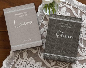 May include: Two personalized wedding vow books. One is light gray with "Laura" and the date "07.25.2025". The other is dark gray with "Oliver" and the same date. Both books have the text "LETTERS & VOWS TO MY LOVE".