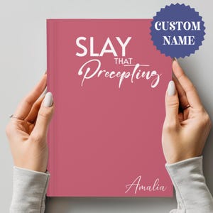 May include: A pink journal with the text "SLAY THAT Precepting" in white. The name "Amalia" is written in white at the bottom of the journal.