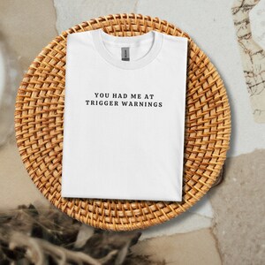 Puede incluir: Camiseta blanca con el texto "YOU HAD ME AT TRIGGER WARNINGS" impreso en negro.