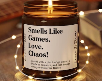 May include: A brown glass candle with a white label that reads "Smells Like Games. Love. Chaos!"  Infused with a pinch of go-getter, a drizzle of romance, and just enough victory to stoke the flames. The candle is lit and sitting on a wooden coaster.