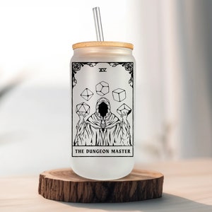 May include: A frosted glass tumbler with a bamboo lid and a straw. The tumbler features a black and white illustration of a figure in a robe holding dice, with the text "The Dungeon Master" and "XV" printed on the glass.