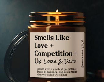 May include: A brown glass candle jar with a white label that reads "Smells Like Love + Competition = Us Lora & David". The label also includes the text "Infused with a pinch of go-getter, a drizzle of romance, and just enough victory to stoke the flames." The candle is lit and the jar has a black lid.