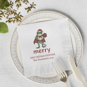 May include: White paper napkin with a festive illustration of a frog wearing a Santa hat and cape, holding a sword and a die. The word "merry" is printed in red, along with "FIRST CRITMAS TOGETHER" and "THE THOMPSONS 2025".