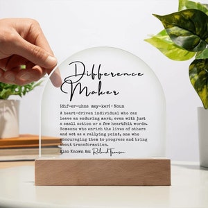 May include: A clear acrylic sign with a rounded top and a wooden base. The sign reads "Difference Maker" with a definition below. The definition reads "[dif-er-uhns mey-ker] Noun A heart-driven individual who can leave an enduring mark, even with just a small action or a few heartfelt words. Someone who enrich the lives of others and act as a rallying point, one who encouraging them to progress and bring about transformation. Also Known As: Roland Tromson"