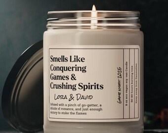 May include: A beige candle with a black lid and a white label that reads "Smells Like Conquering Games & Crushing Spirits Lora & David" and "Infused with a pinch of go-getter, a drizzle of romance, and just enough victory to stoke the flames". The label also reads "Game Night 2025", "9 oz eco vegan soy wax candle", "50-60 hours burning time", and "Keep away from flammables".